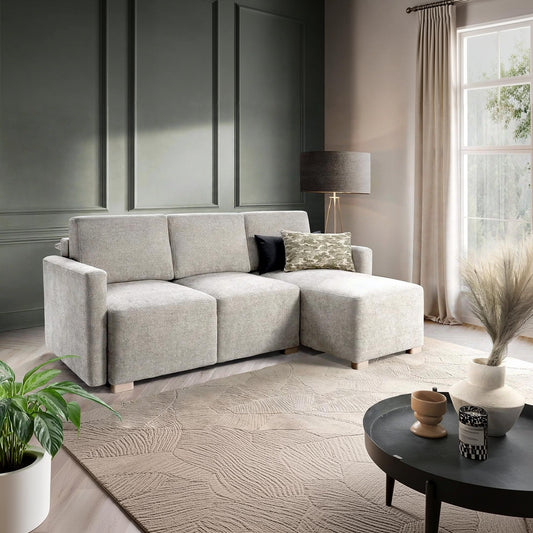 A light grey upholstered sectional sofa with a chaise lounge, styled in a modern living room with a textured beige rug, a dark round coffee table, and a window with beige curtains.