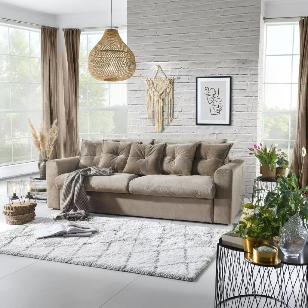 A beige fabric sofa with matching throw pillows in a bright, modern living room. The room features a white brick wall, a woven pendant light, a macrame wall hanging, and a black-framed abstract art print.
