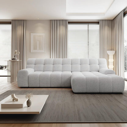 A modern light gray tufted sectional sofa in a bright, contemporary living room with beige curtains, a gray rug, and a low white coffee table.
