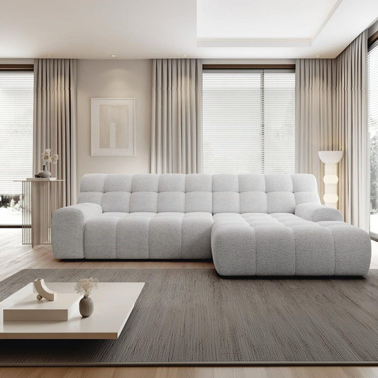 A modern light gray tufted sectional sofa in a bright, contemporary living room with beige curtains, a gray rug, and a low white coffee table.